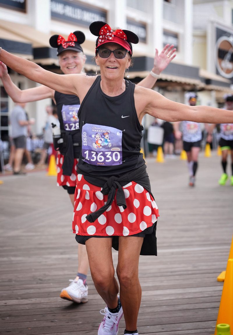 Paula running in Minnie Mouse ears at a Disney marathon, arms thrown wide, bib reading Paula.
