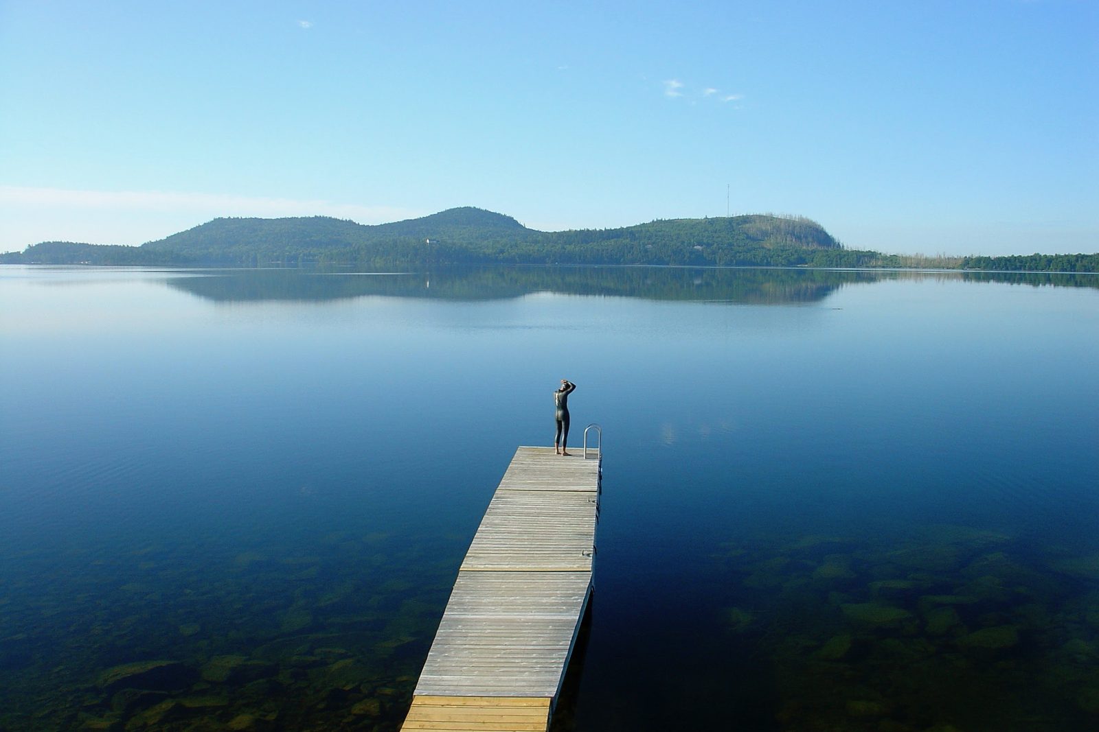 A swimmer in a wetsuit stands at the end of a long dock on a glass-still lake at morning, forested hills on the horizon.