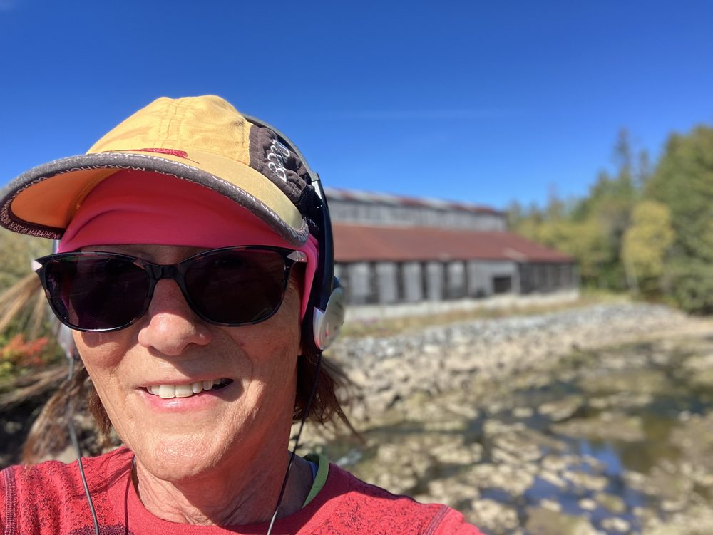 Close selfie of Paula in a Boston Marathon cap on a bright morning beside a rocky river.